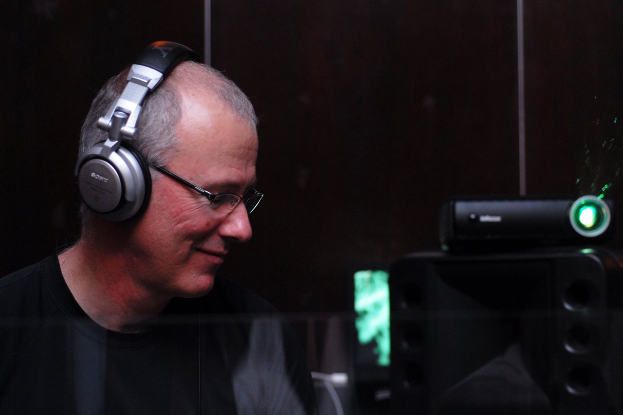 A DJ wearing headphones and glasses smiles while performing, with speakers and equipment in the background.