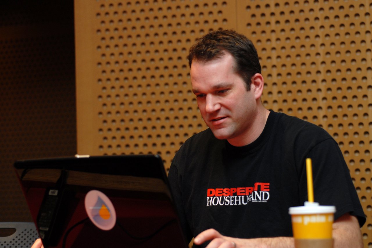 A man in a black t-shirt is working on a laptop in a conference setting, with a drink nearby.