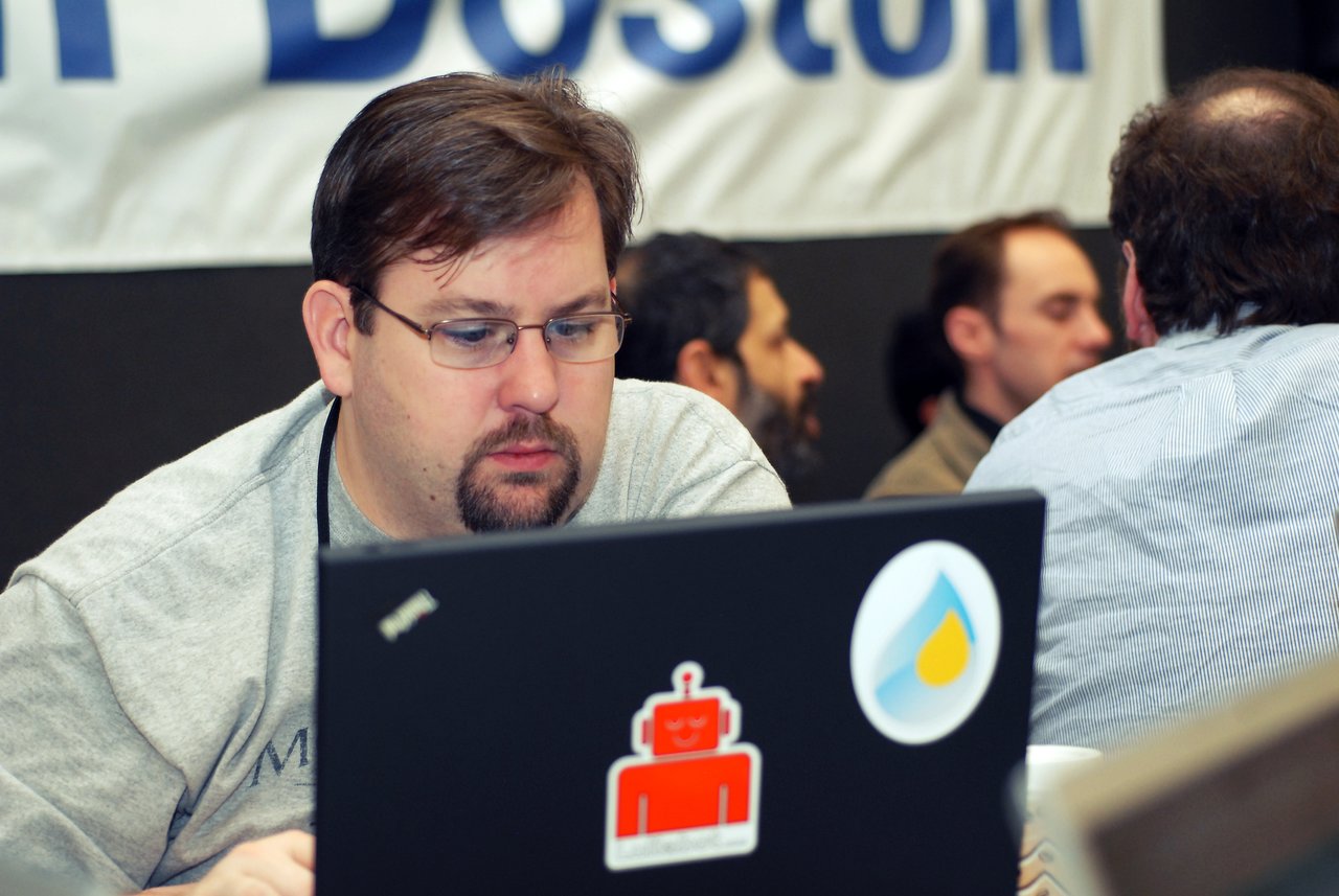 A man wearing glasses and a gray shirt focuses on his laptop at a conference, with others in the background.