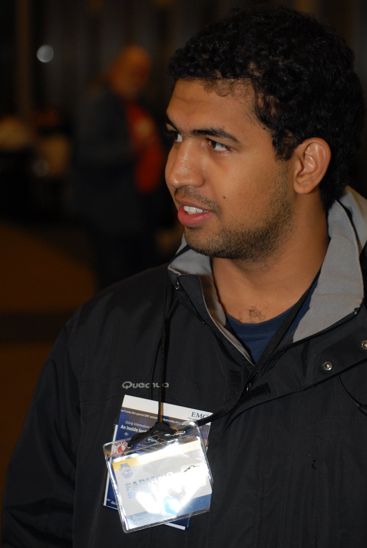 A man wearing a conference badge and a black jacket is engaged in conversation at an event.