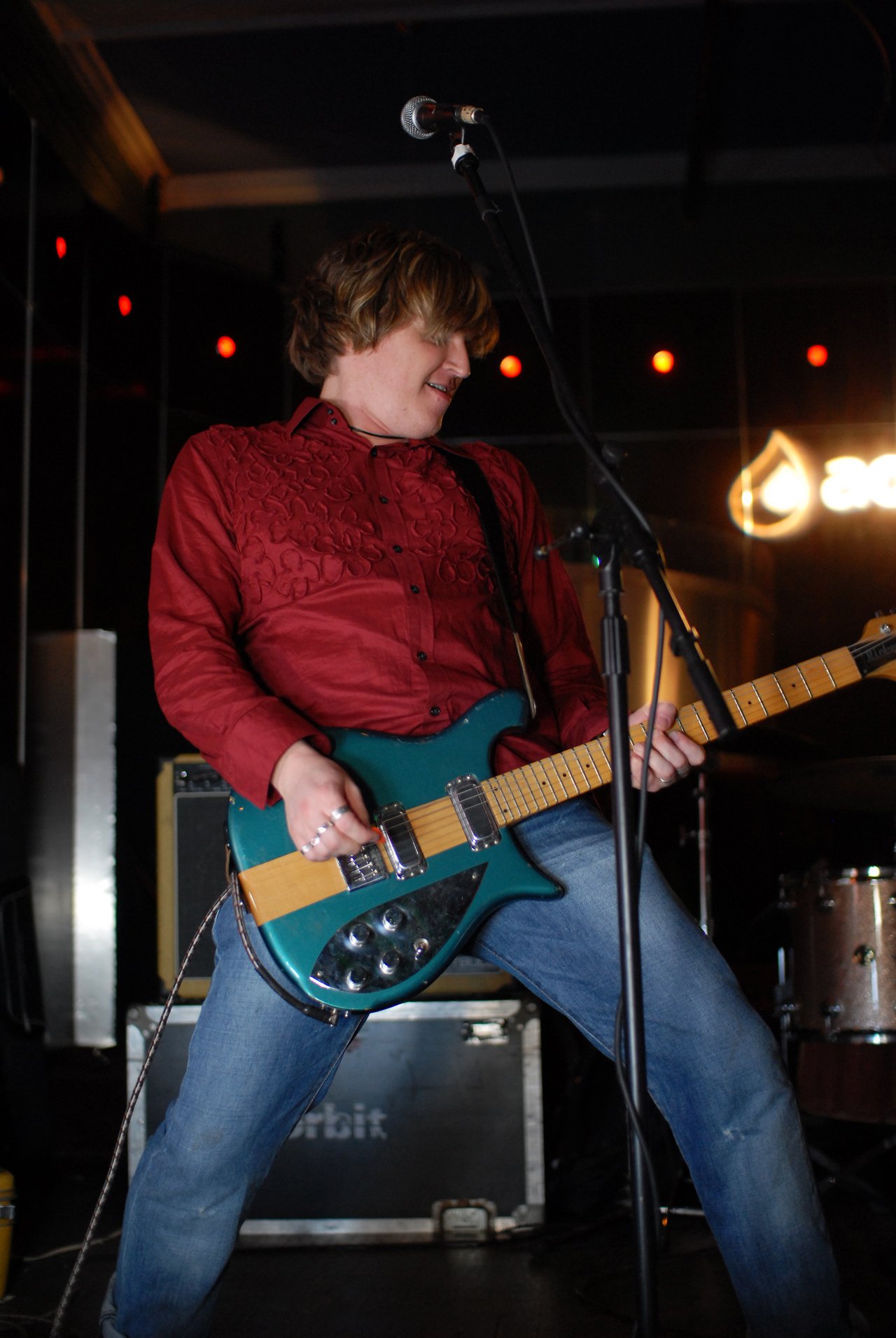 A musician in a red shirt plays an electric guitar on stage, looking engaged in the performance.