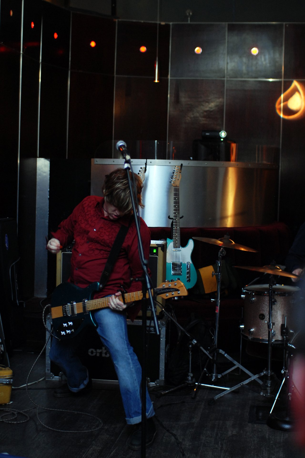 A guitarist in a red shirt plays an electric guitar energetically on stage, with drums and other instruments in the background.