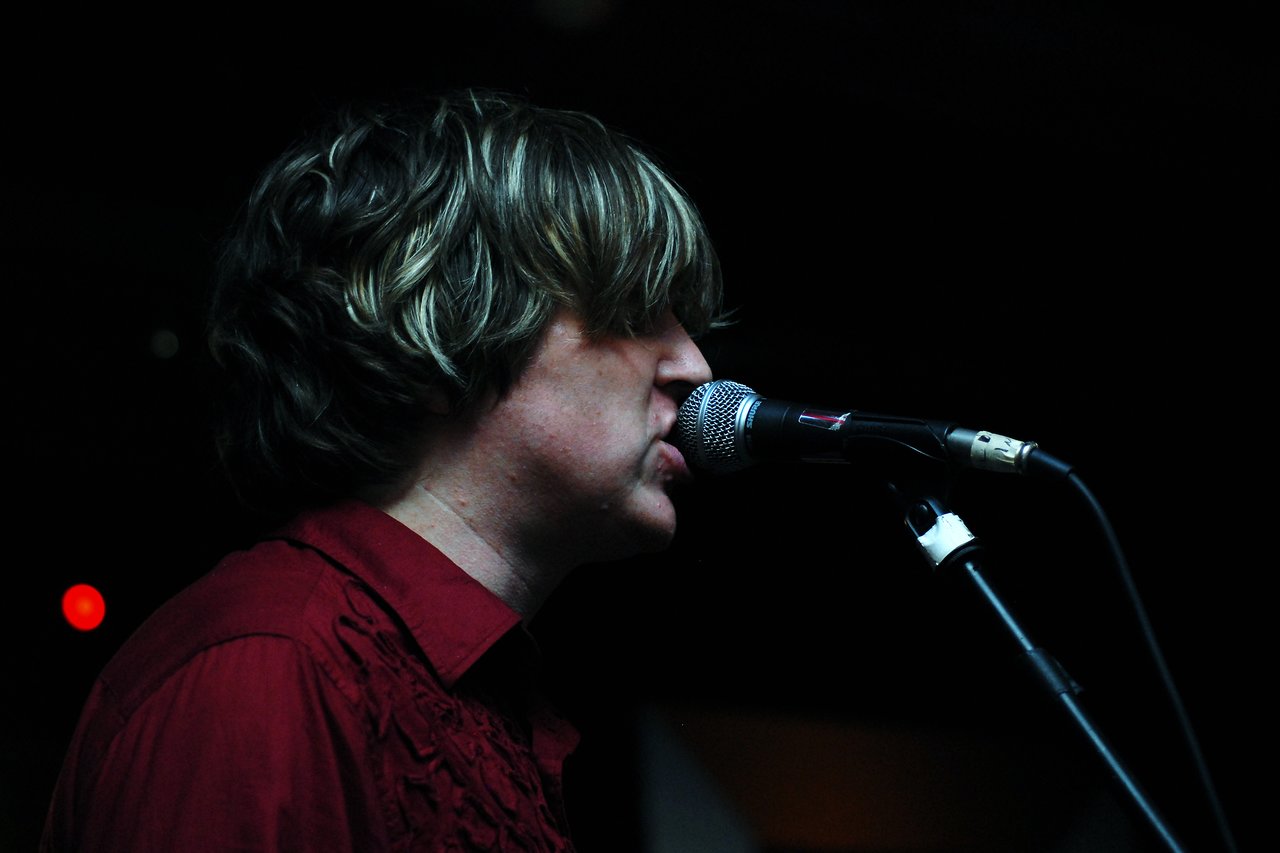 A person in a red shirt sings into a microphone on stage, with a dark background.
