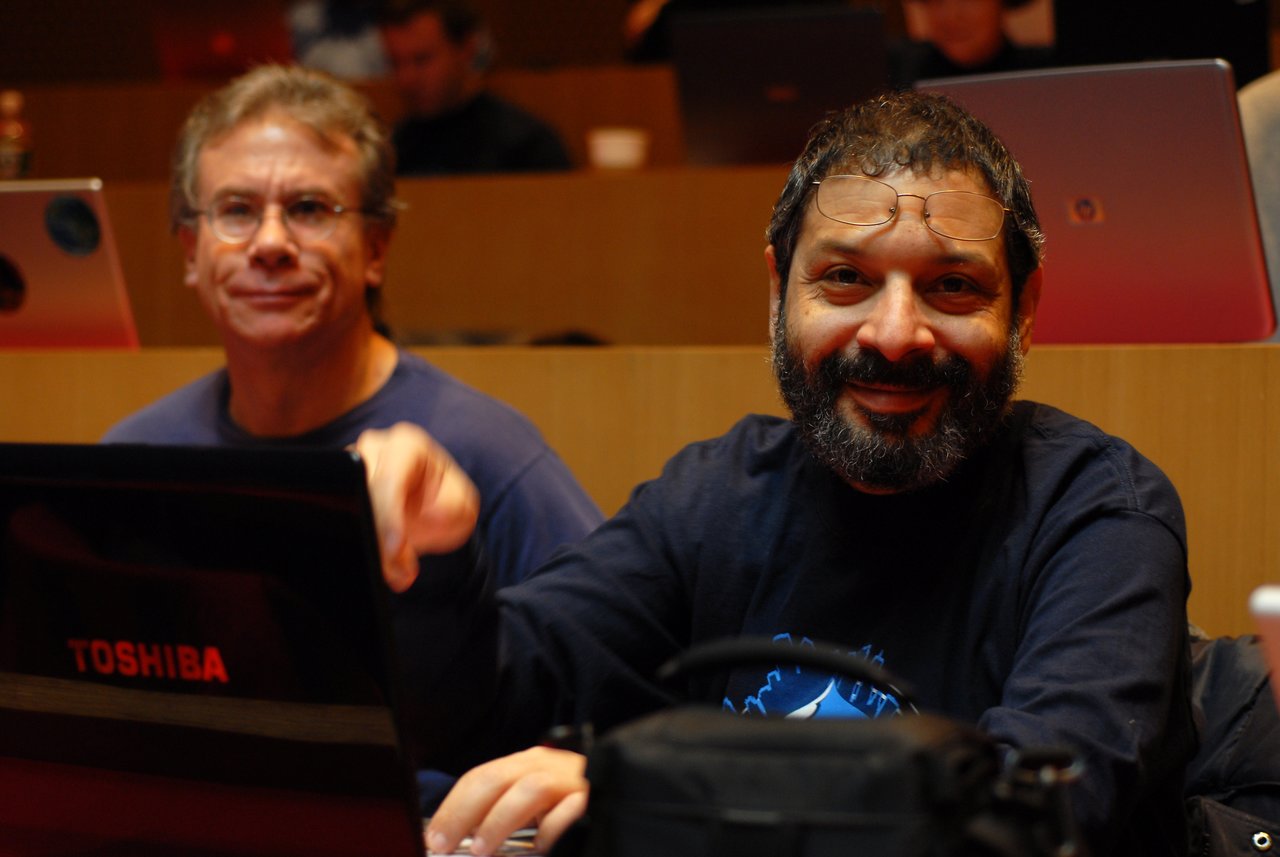 Two men sitting in a conference room with laptops, one smiling at the camera while the other looks focused.