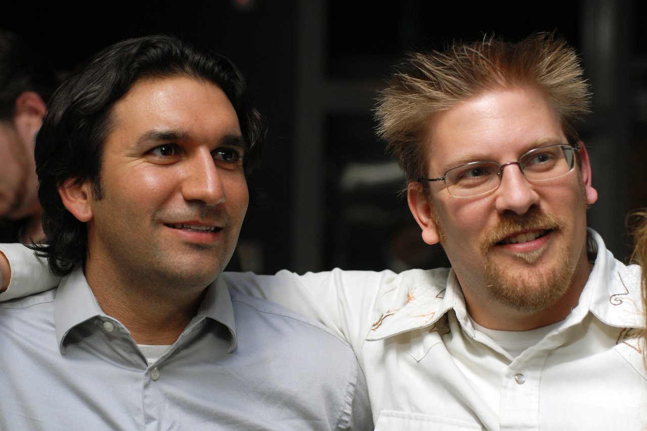 Two men smiling, one with his arm around the other, at a social event.