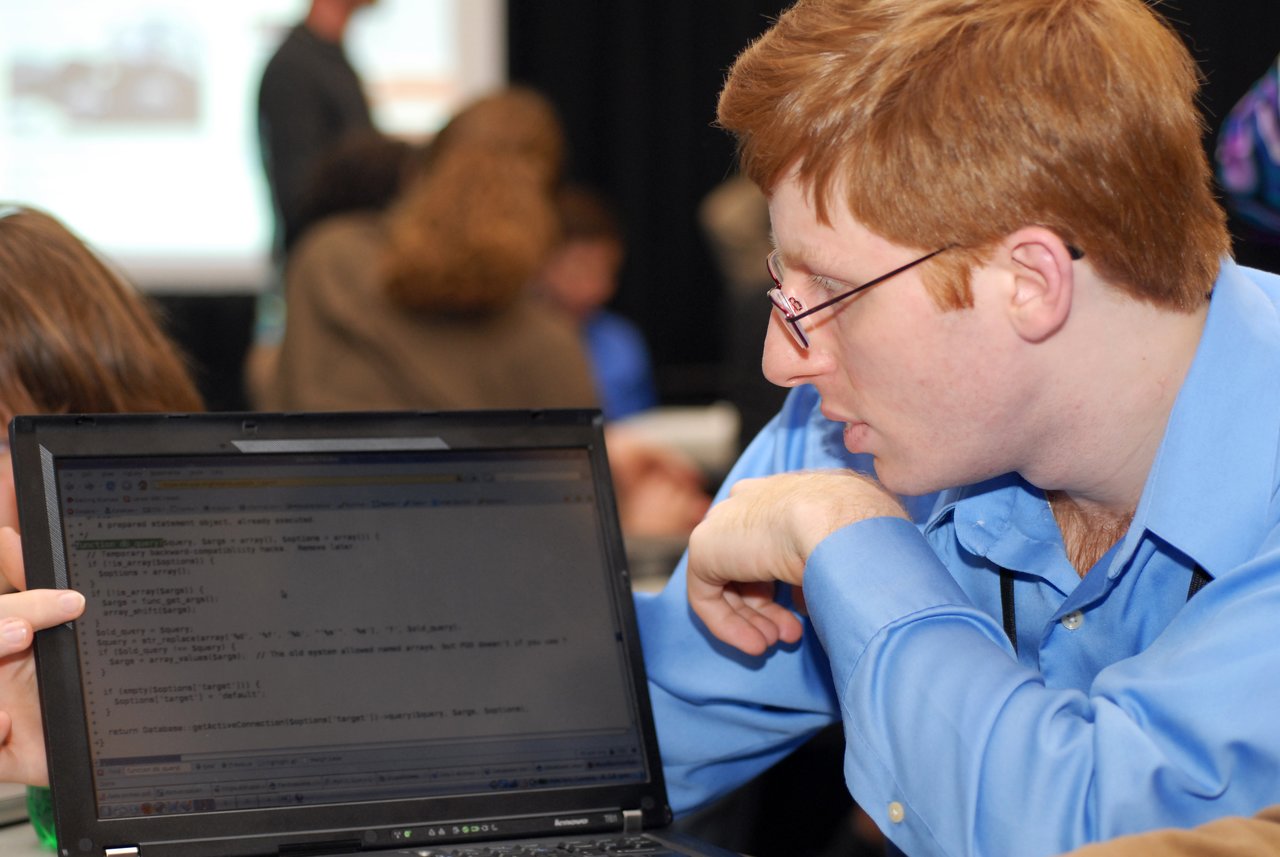 A person in a blue shirt examines a laptop screen displaying code, discussing a Drupal 7 database improvement.