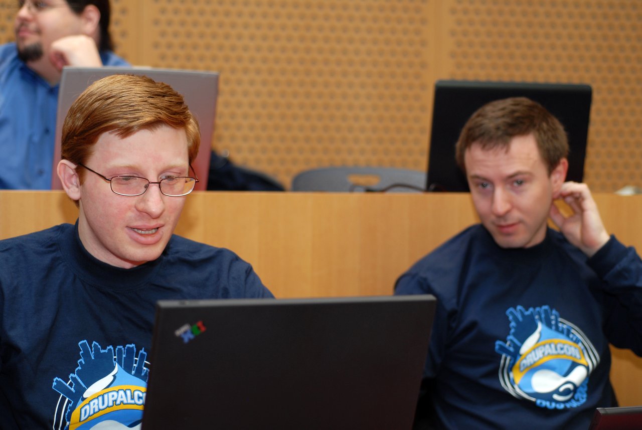 Two people wearing DrupalCon shirts sit in a conference room, one focused on a laptop while the other listens.
