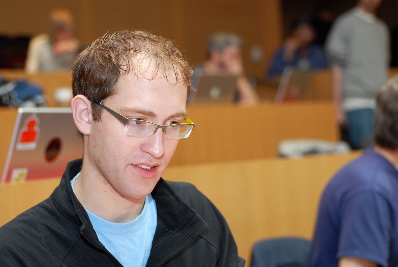 A man wearing glasses and a black jacket is focused on something in front of him at a conference.