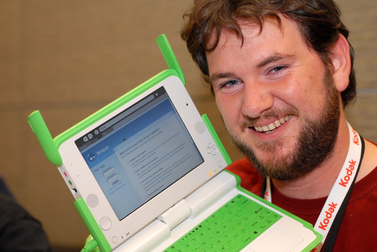 A person smiles while holding up an OLPC laptop displaying a Drupal website on its screen.