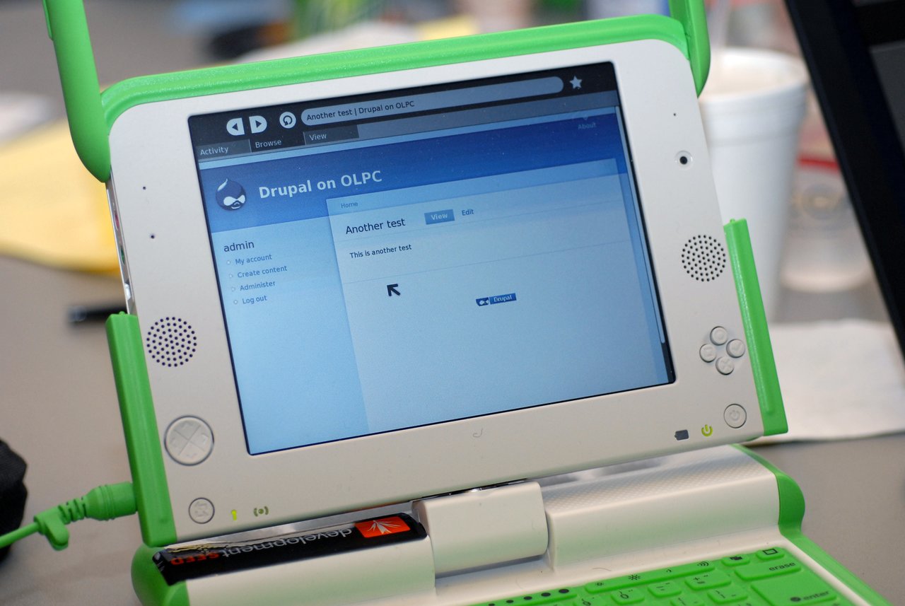 A green and white OLPC laptop displays a Drupal website on its screen, with a USB drive plugged in.