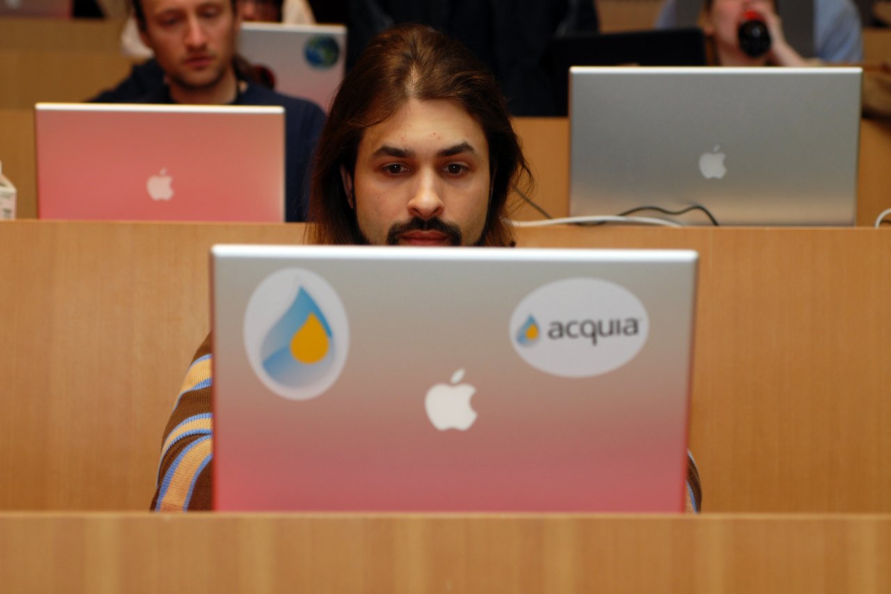 A person with long hair and a beard is focused on a laptop with Acquia stickers in a conference setting.