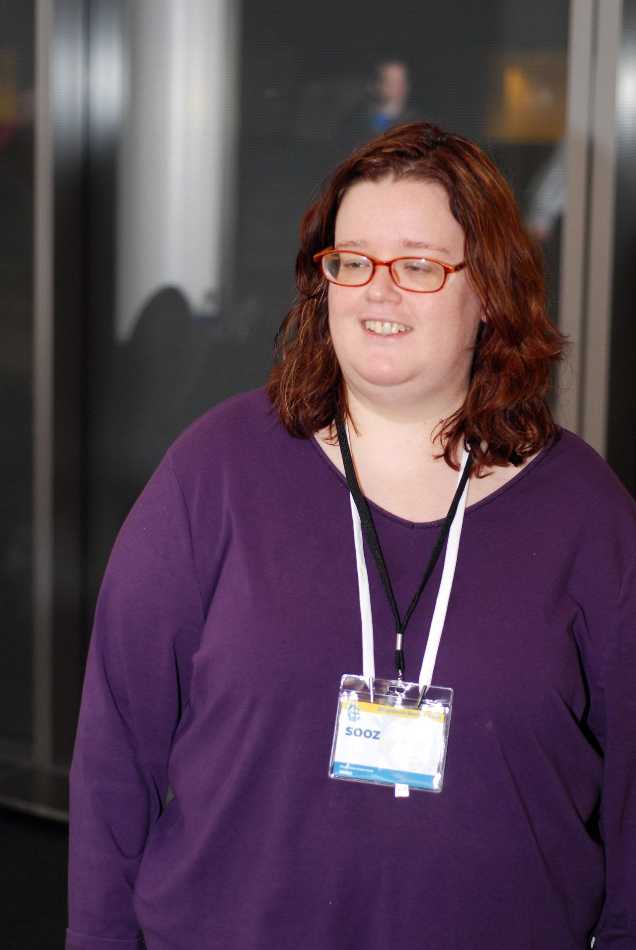 A person wearing glasses and a purple shirt stands indoors with a conference badge labeled "Sooz" around their neck.