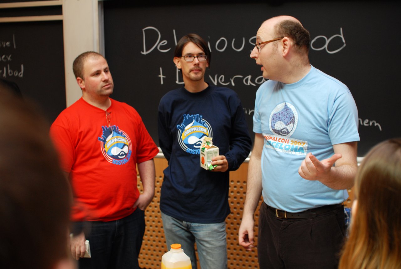 Three people in DrupalCon shirts stand together, one gesturing while speaking, as others listen in a discussion.