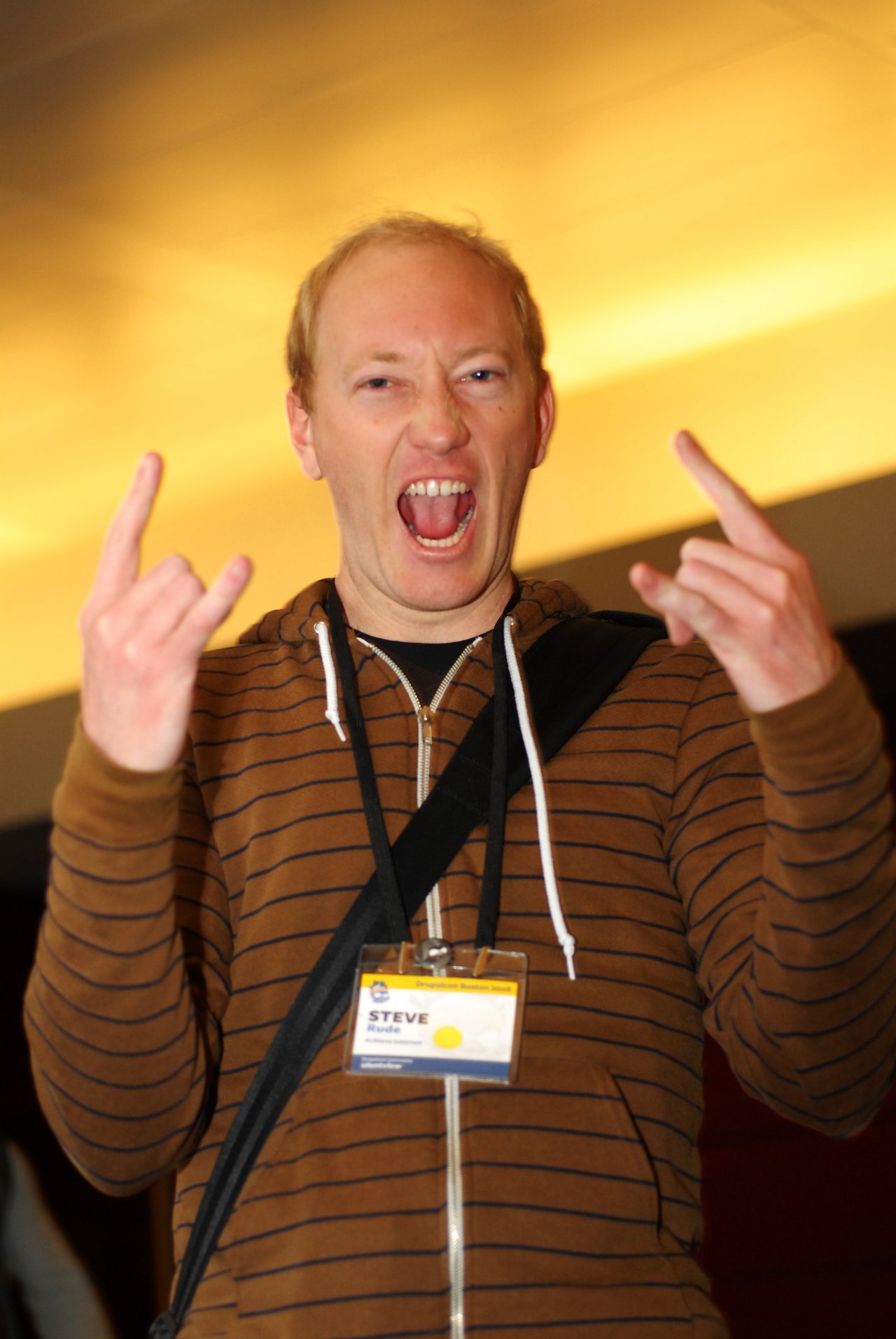 A person wearing a striped hoodie and a name badge makes a rock hand gesture and smiles energetically.