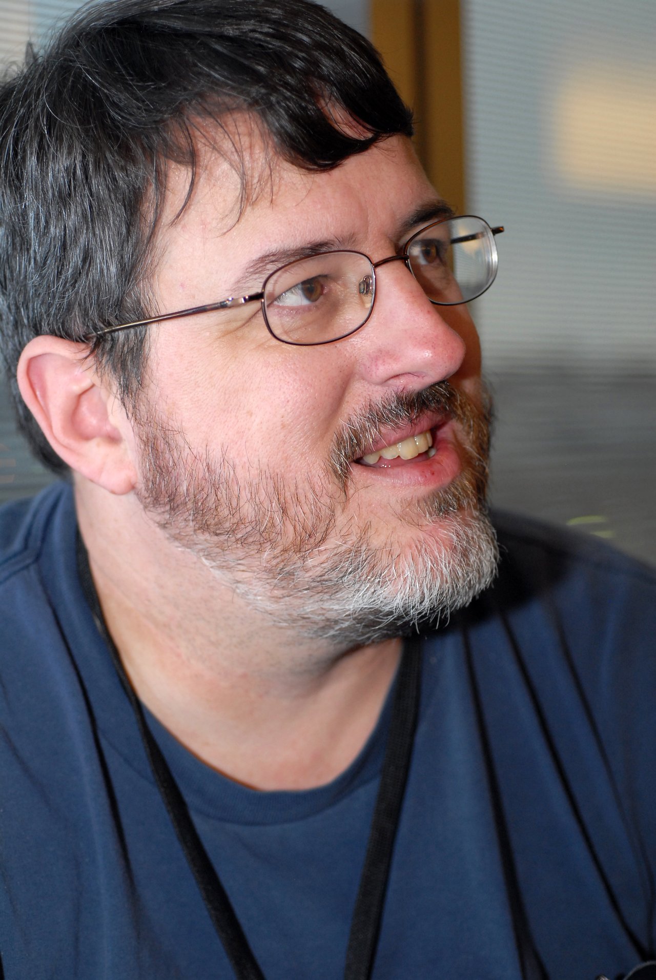 A man with glasses and a beard wearing a blue shirt looks to the side and smiles.