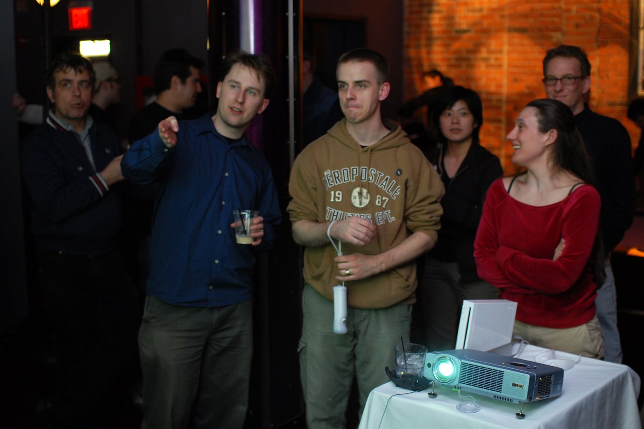 A group watches as a man holds a Wii controller, appearing focused.