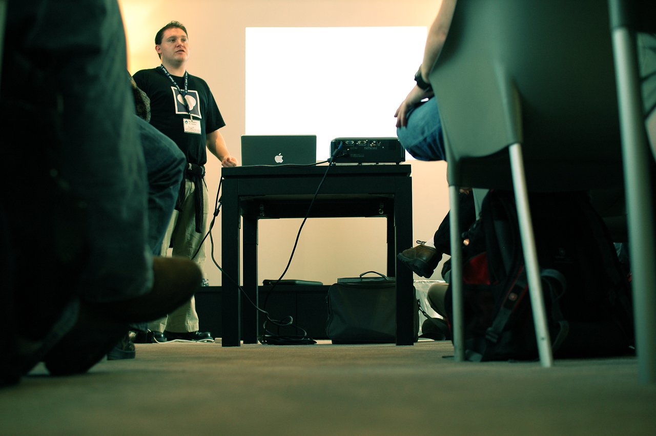 A presenter stands at the front of a room, speaking to an audience with a laptop and projector.
