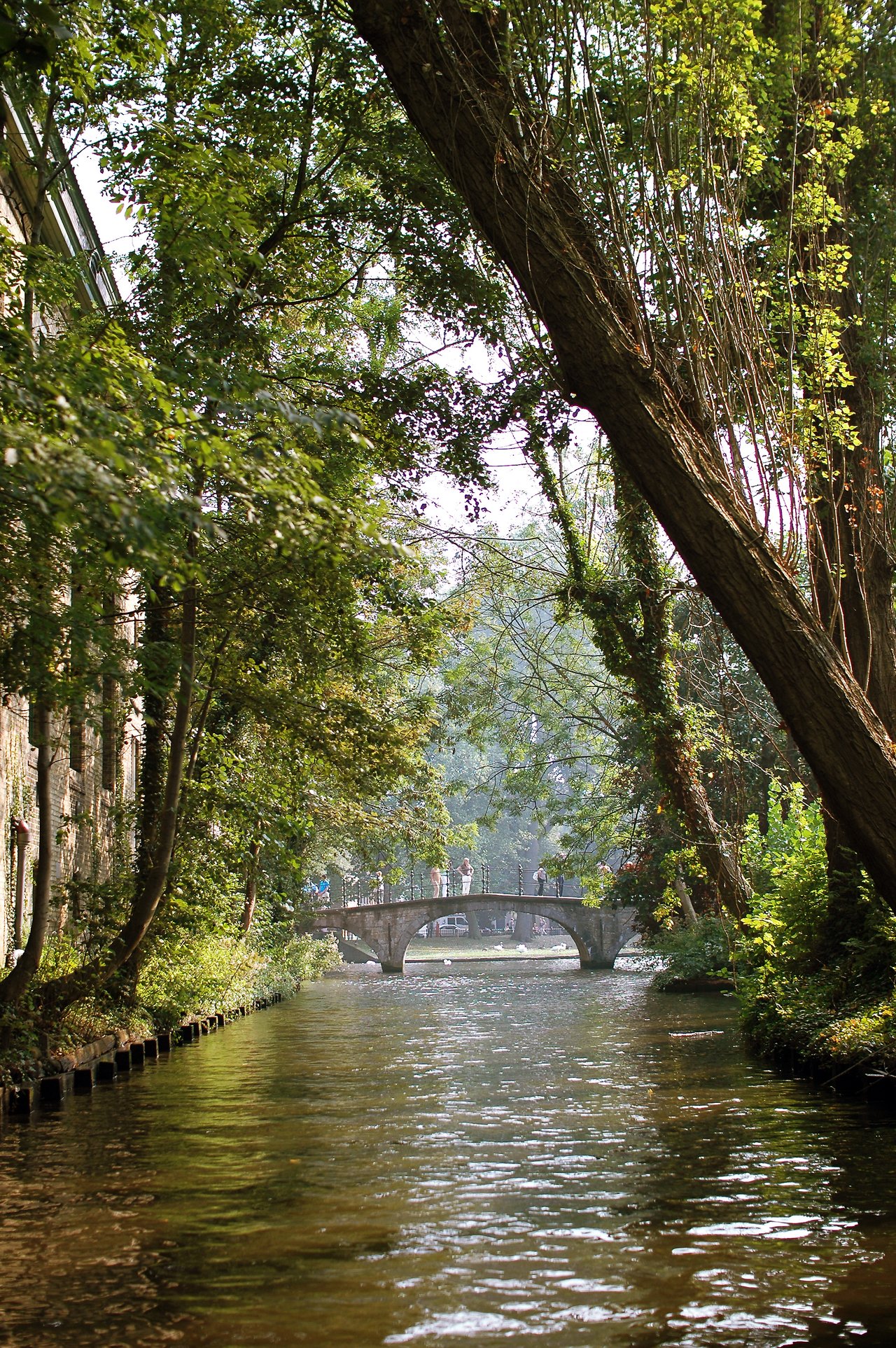A canal in Bruges with a stone bridge, surrounded by green trees, and people walking across.