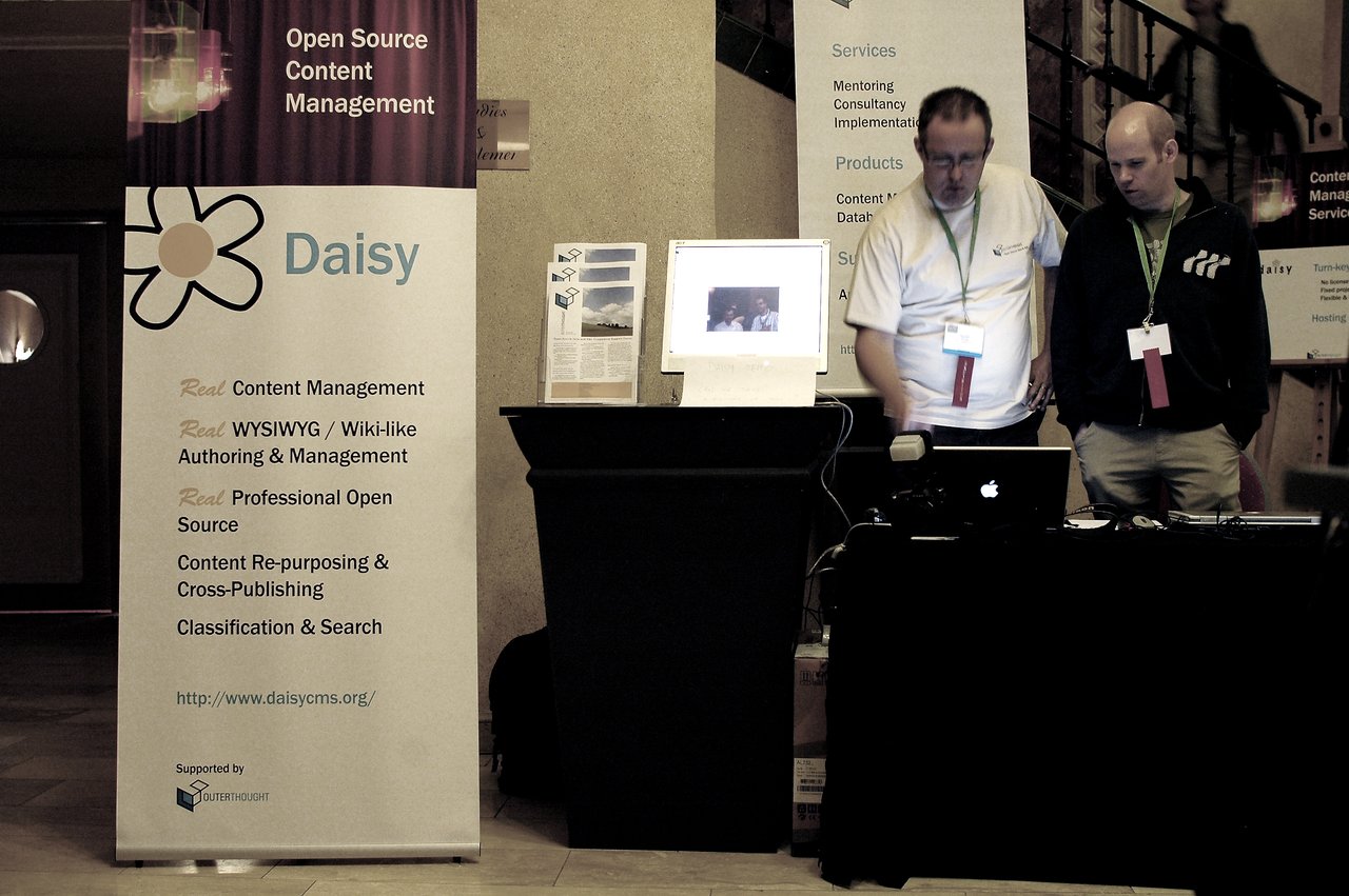 Two men stand at a booth with a "Daisy" banner, looking at a laptop screen during DrupalCon Brussels 2006.