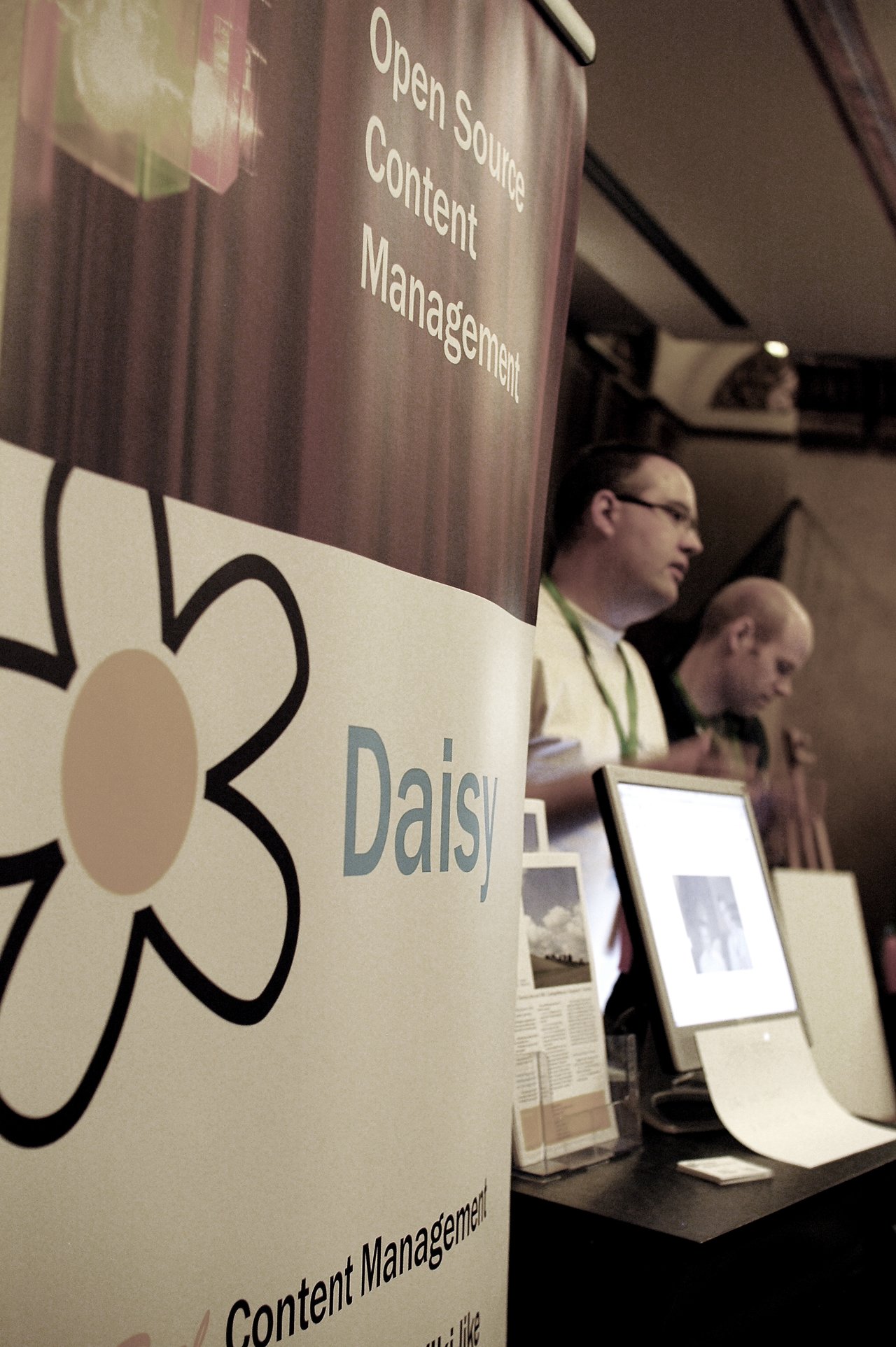 A booth at DrupalCon Brussels 2006 promoting "Daisy" open-source content management, with two people discussing near a computer.