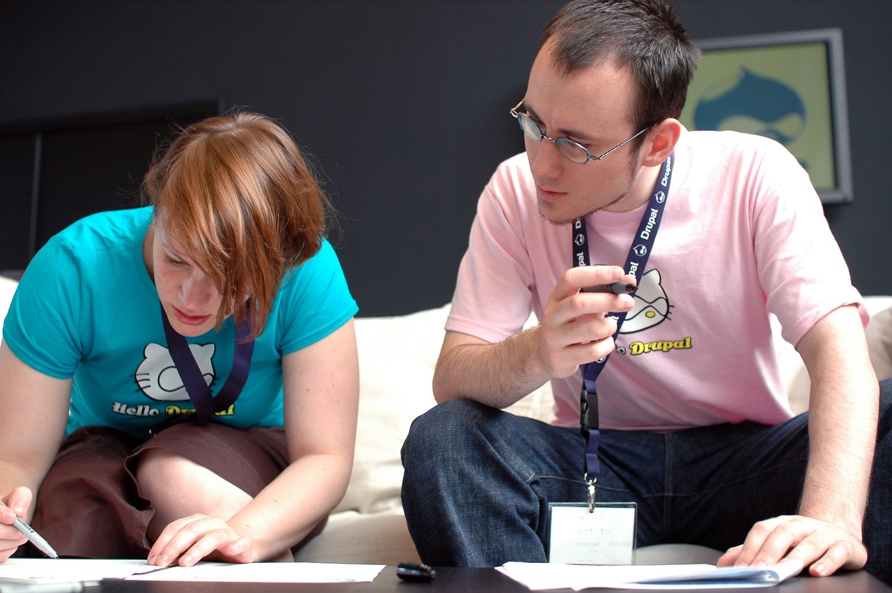 Two people in Drupal-themed shirts are sitting together, writing on papers and discussing their work.