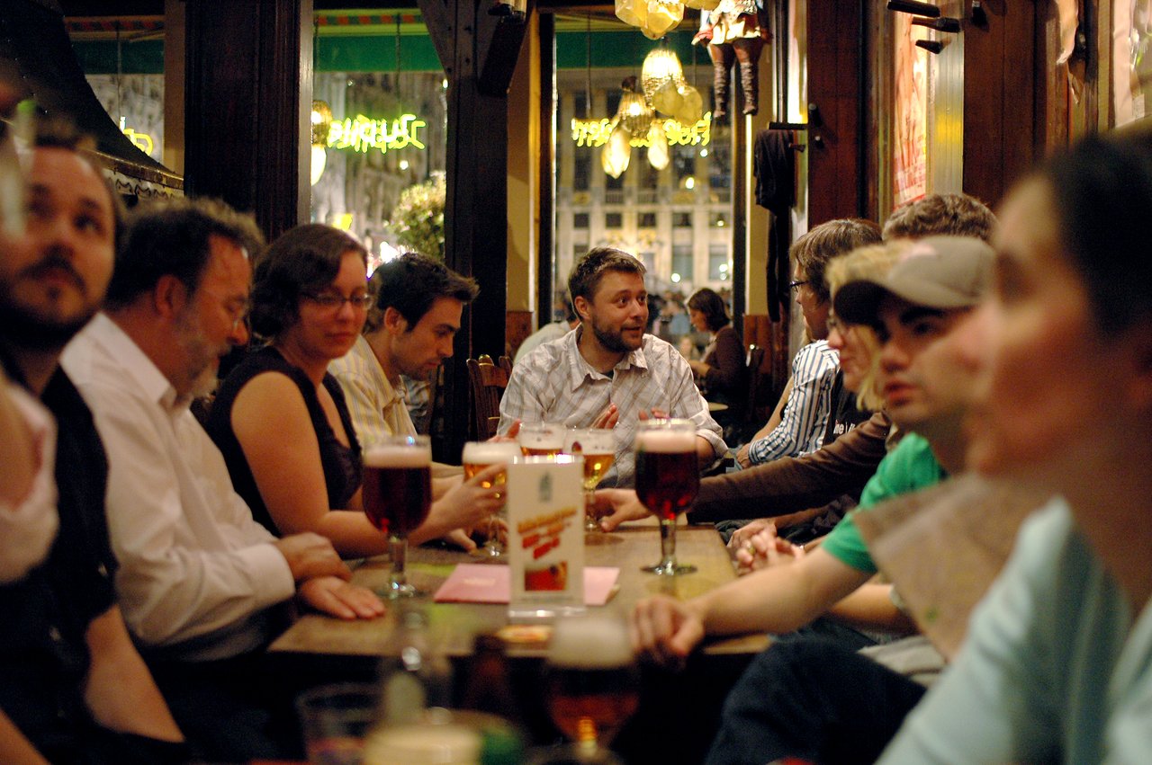 A man in a plaid shirt speaks while seated at a table with drinks and a group of people.