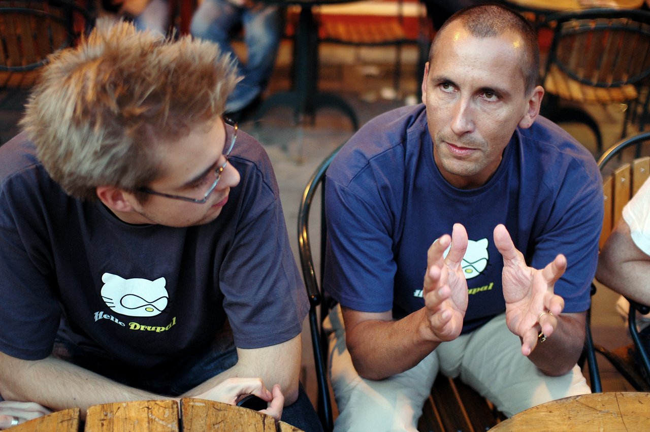 Two people in matching Drupal shirts sit at a table, one gesturing while speaking and the other listening attentively.