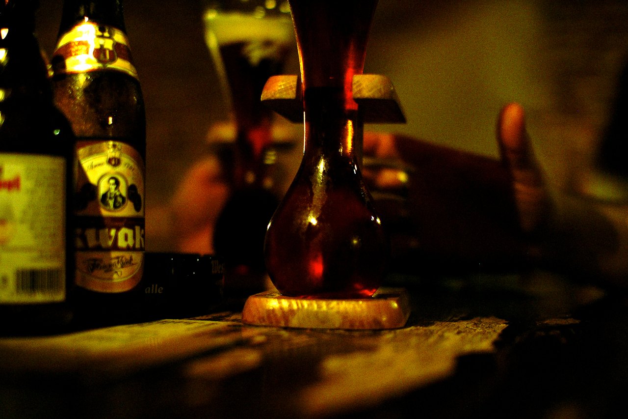 A Kwak beer in its signature glass with a wooden stand, surrounded by bottles and hands holding drinks.