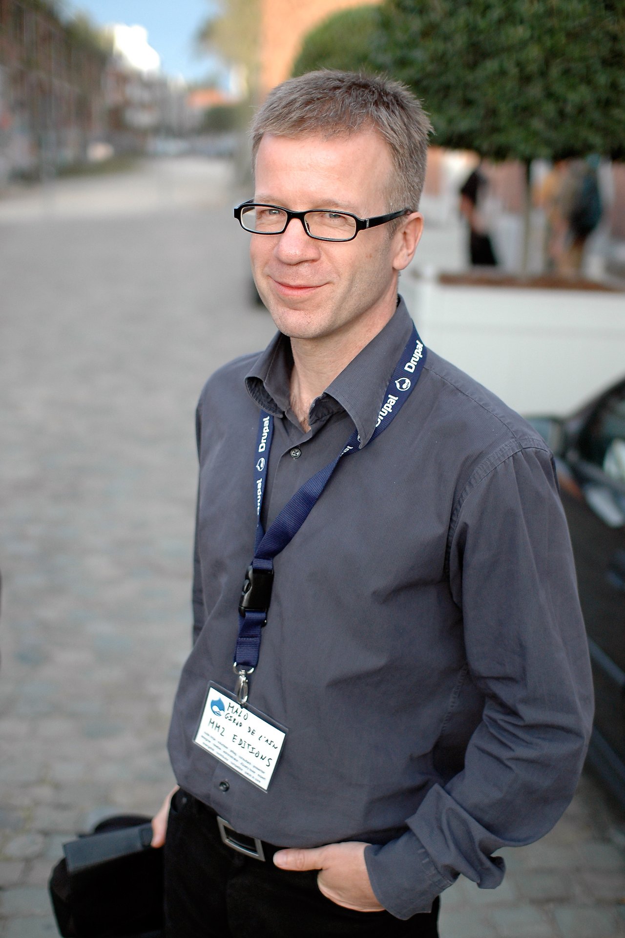 A man wearing glasses and a Drupal lanyard stands outdoors, smiling slightly at the camera.