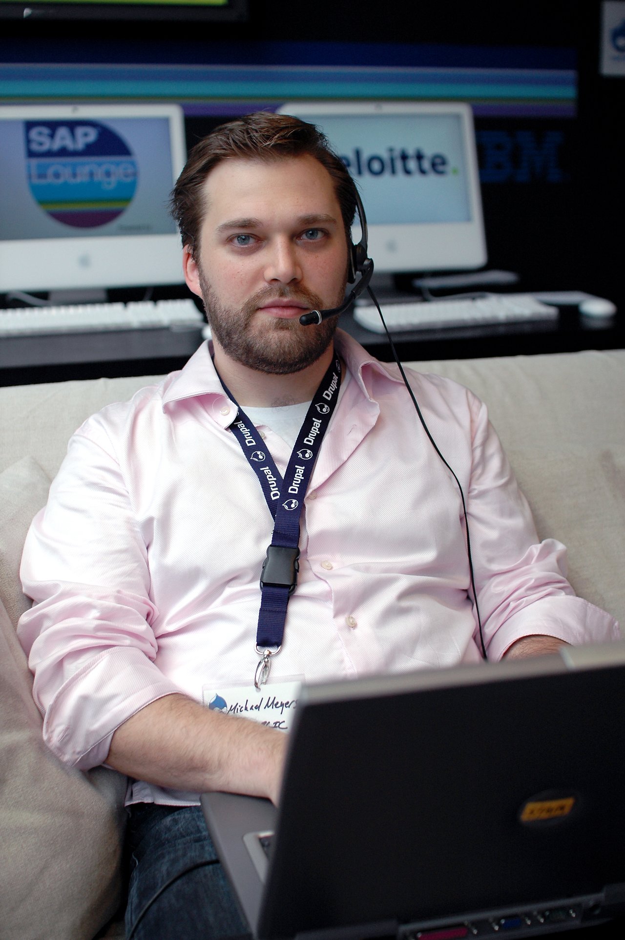 A man wearing a headset and Drupal lanyard sits on a couch using a laptop at a tech event.