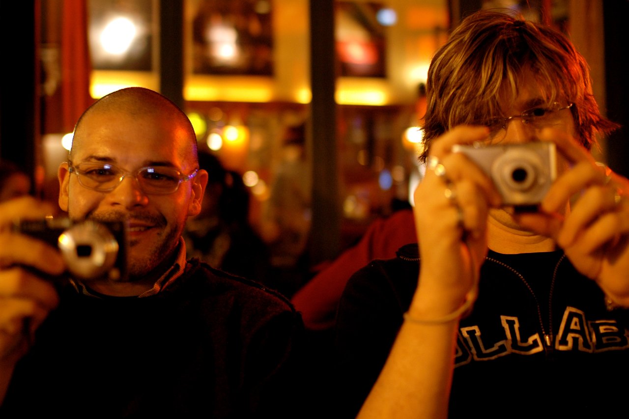 Two people in a dimly lit setting smile while holding cameras and taking photos.