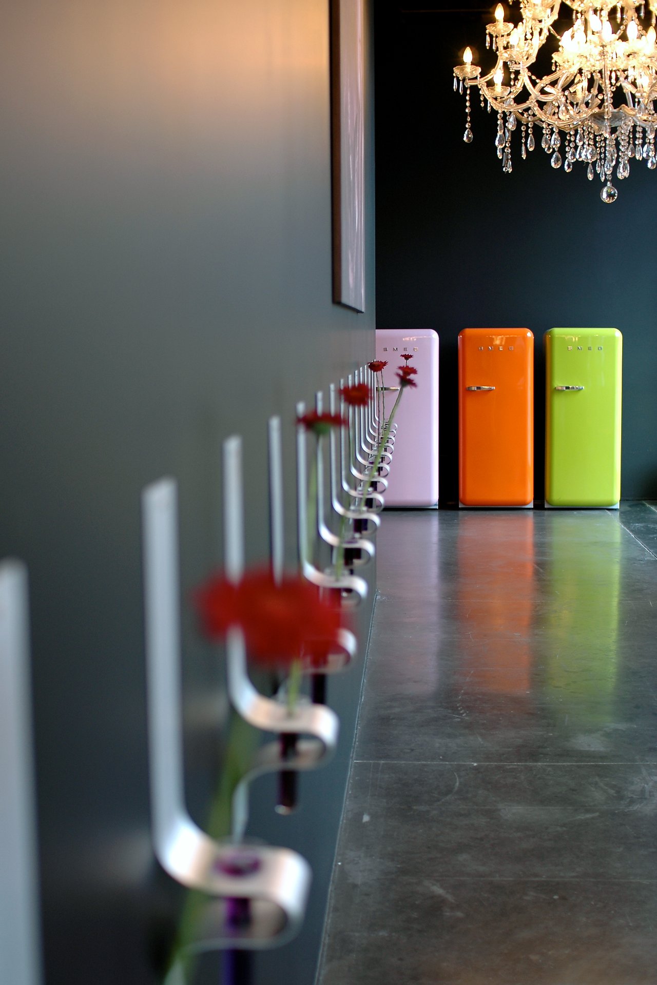 A row of red flowers in wall-mounted vases leads to three colorful refrigerators under a chandelier.