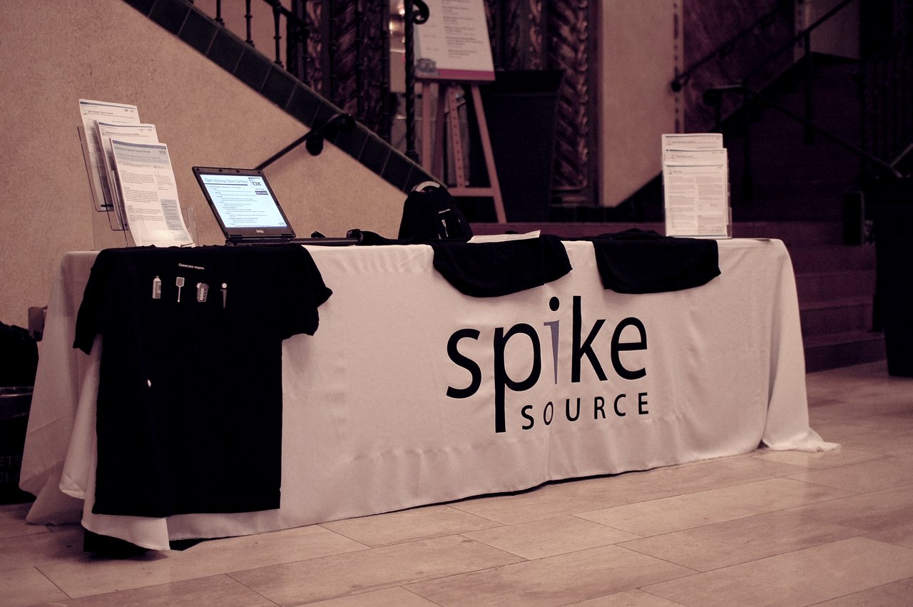 A SpikeSource exhibitor table displaying promotional materials, a laptop, printed documents, and black t-shirts at a conference.