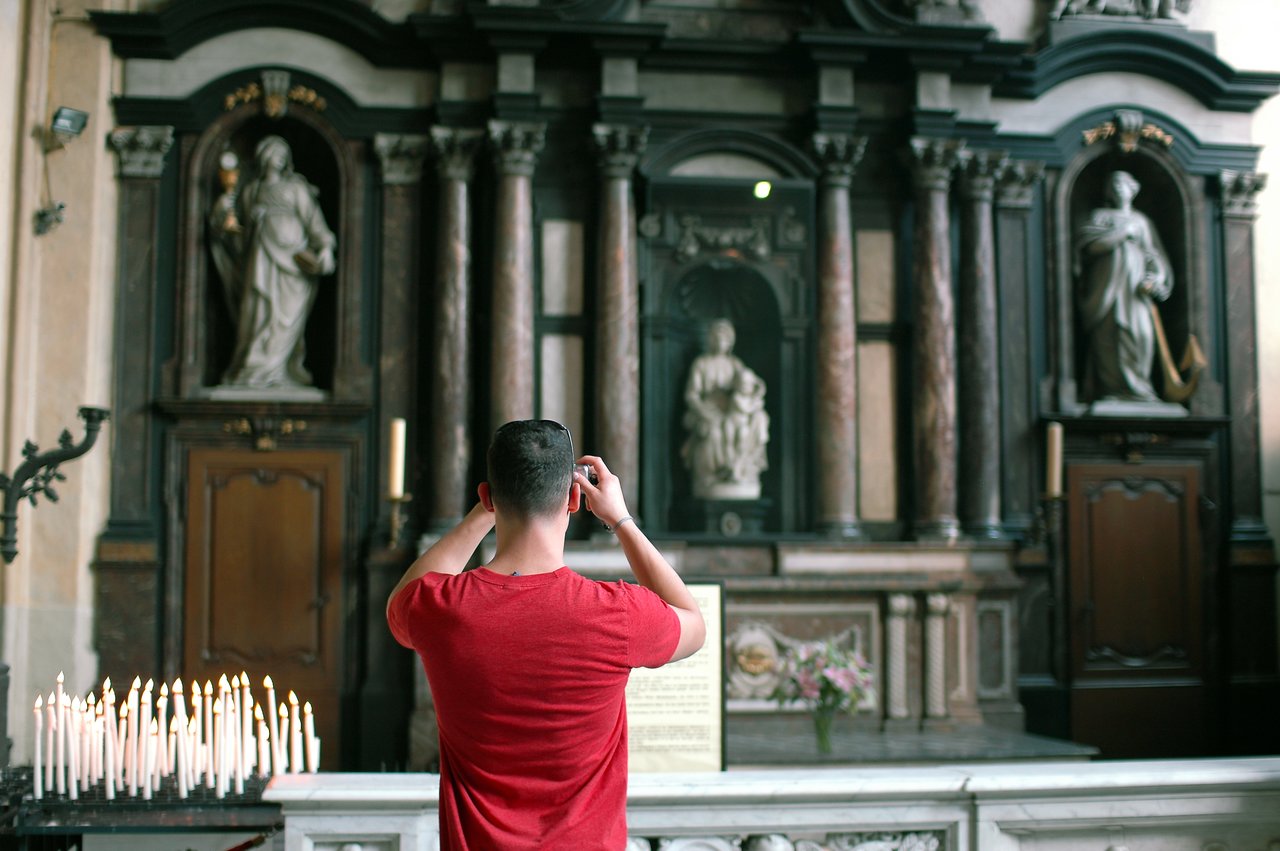 A person in a red shirt takes a photo of Michelangelo's Madonna and Child statue inside a church.