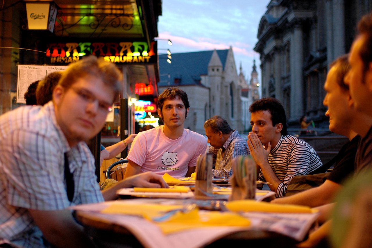 A group of people sits at an outdoor restaurant table, with one person wearing a pink "Hello Drupal" shirt.