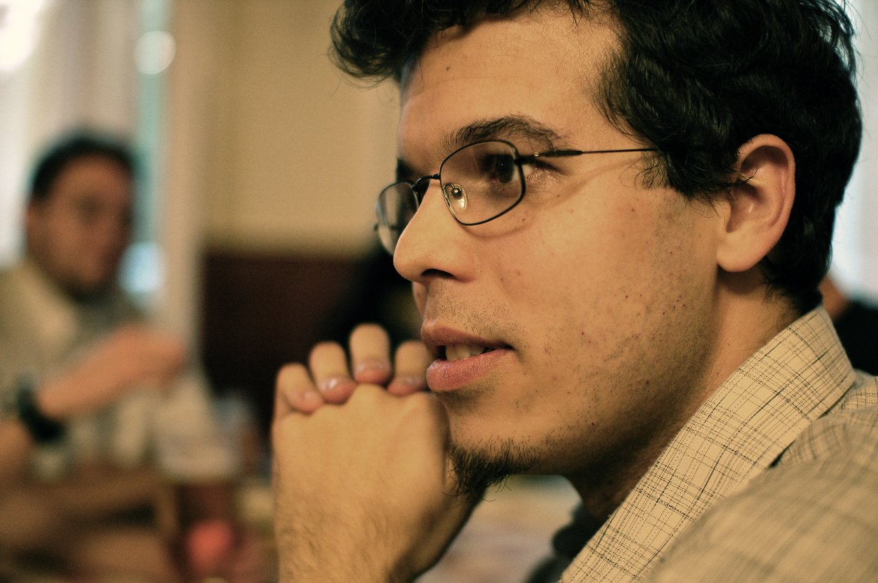 A man wearing glasses and a plaid shirt sits with hands clasped, attentively listening in a social setting.