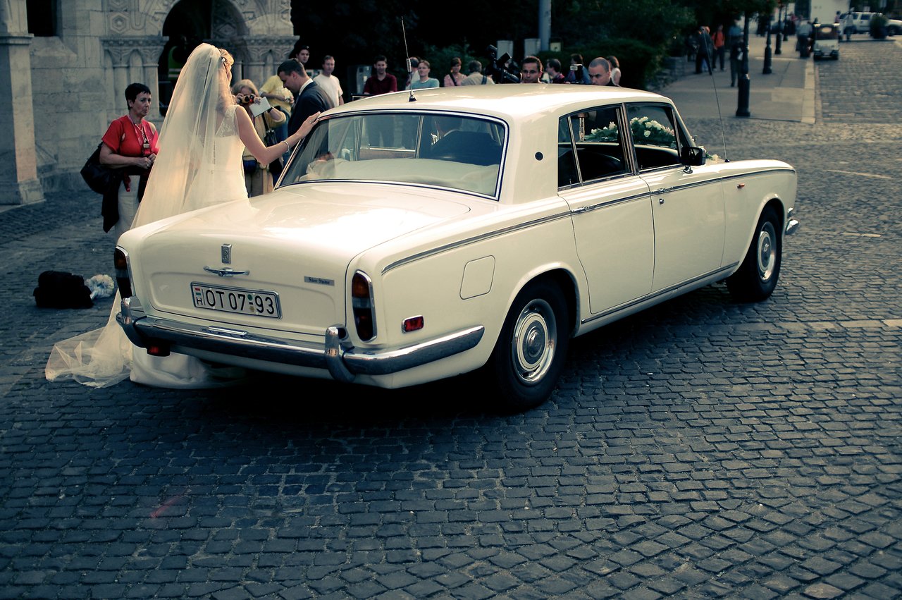 A bride in a white gown stands beside a classic white car, preparing to enter as people watch nearby.