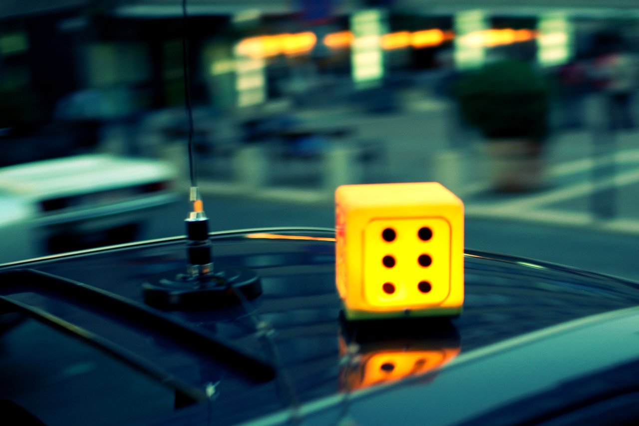 A taxi with a glowing yellow dice-shaped sign on its roof waits on a busy city street.