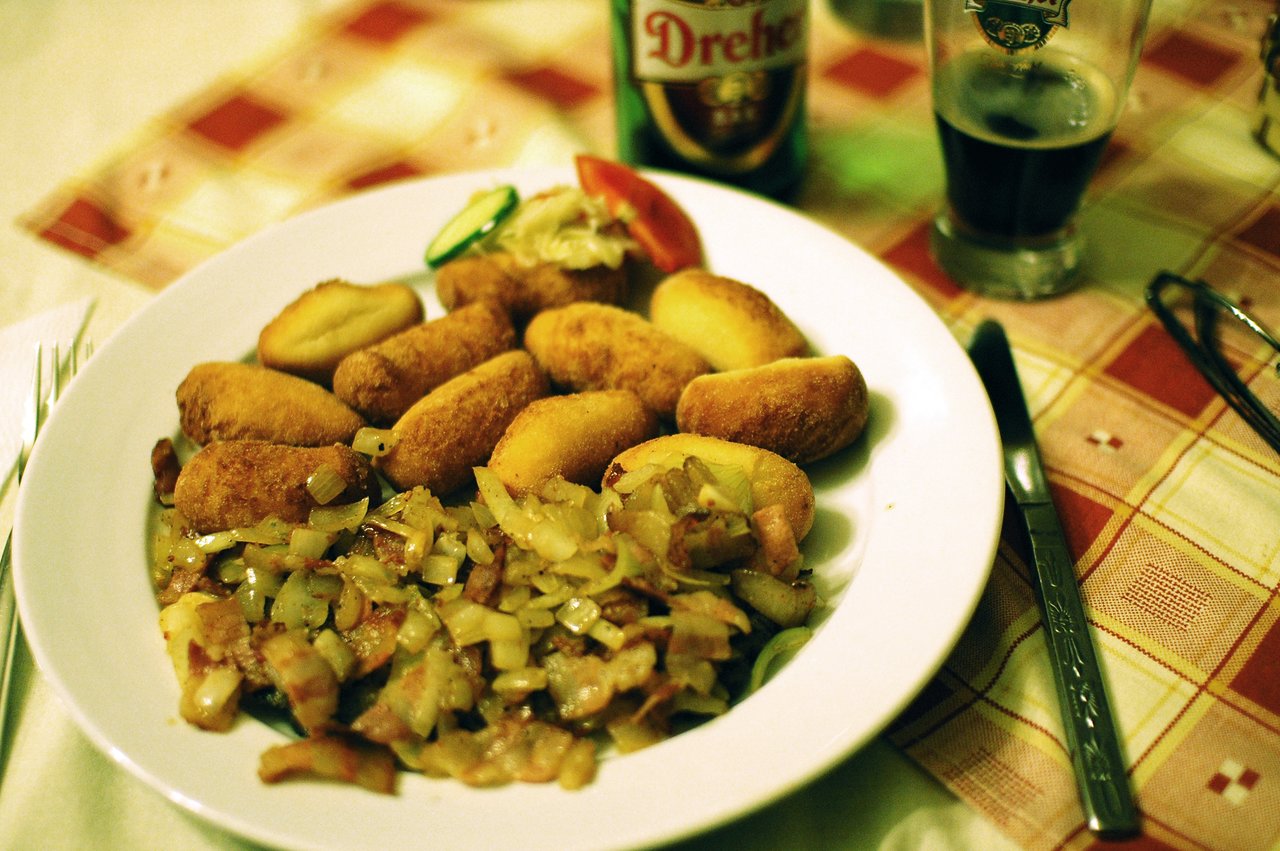 A plate of fried potato croquettes with sautéed onions and bacon, served with vegetables and drinks on a table.
