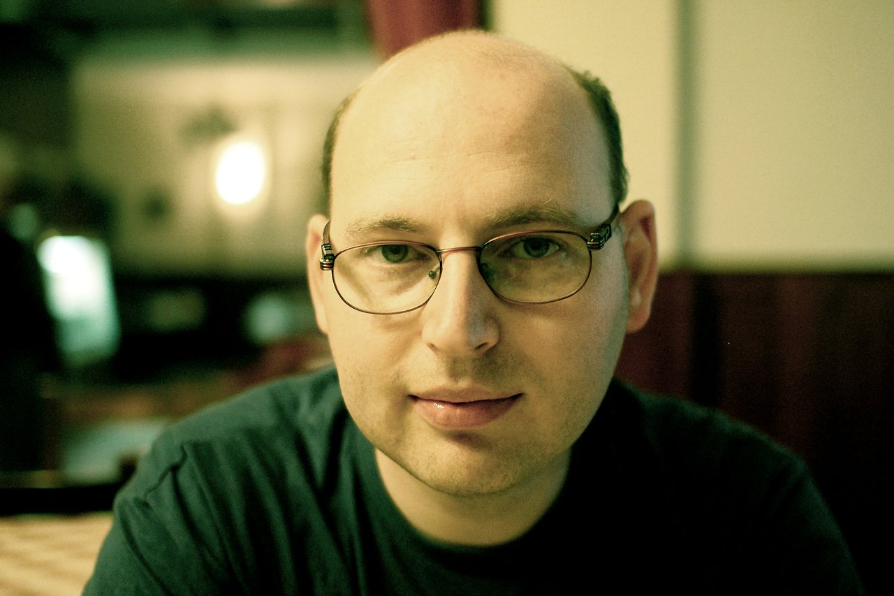 A man with glasses and a dark shirt looks directly at the camera in a dimly lit indoor setting.