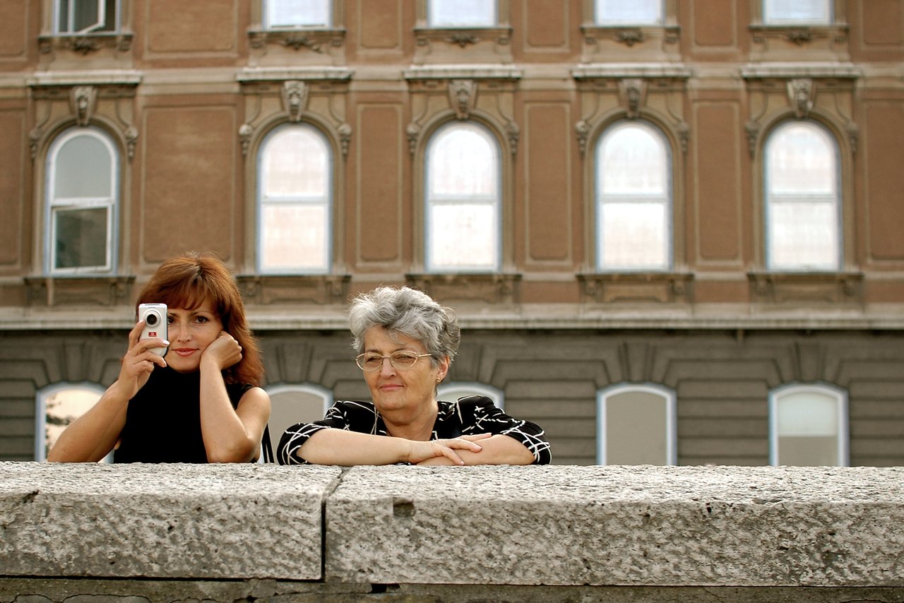 A woman holds a small camera while another woman leans on a stone ledge, both looking in different directions.