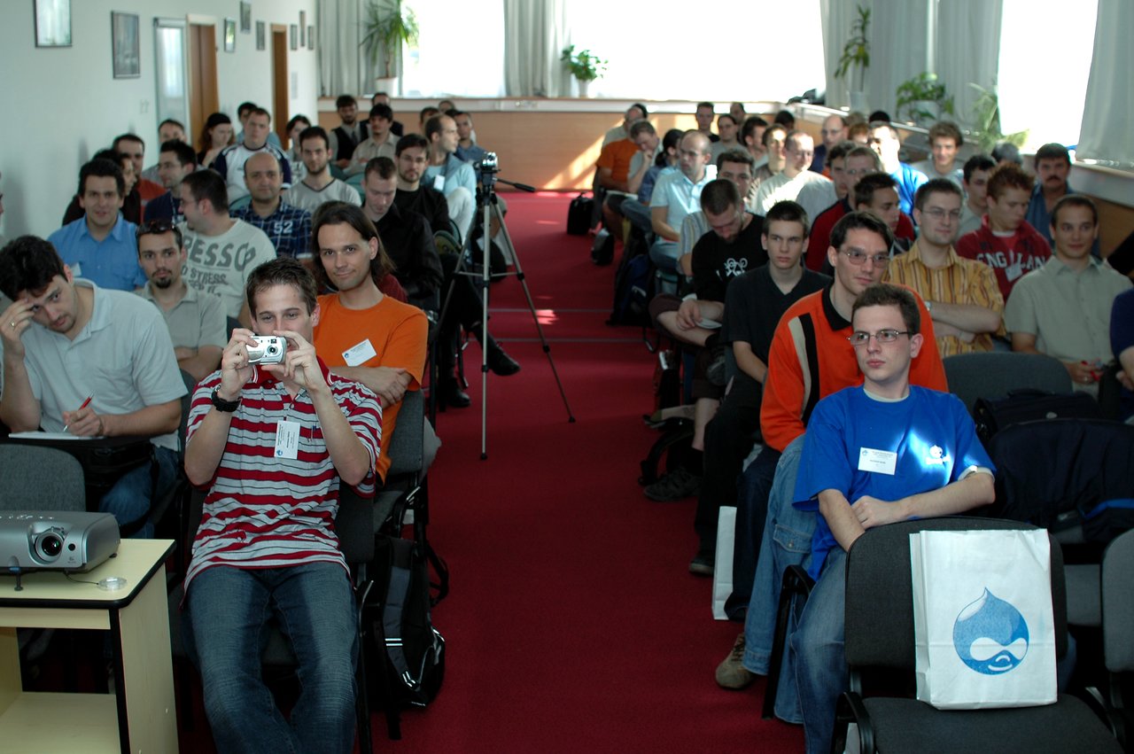 A full conference room with attendees seated, some taking notes, while one person in the front takes a photo.