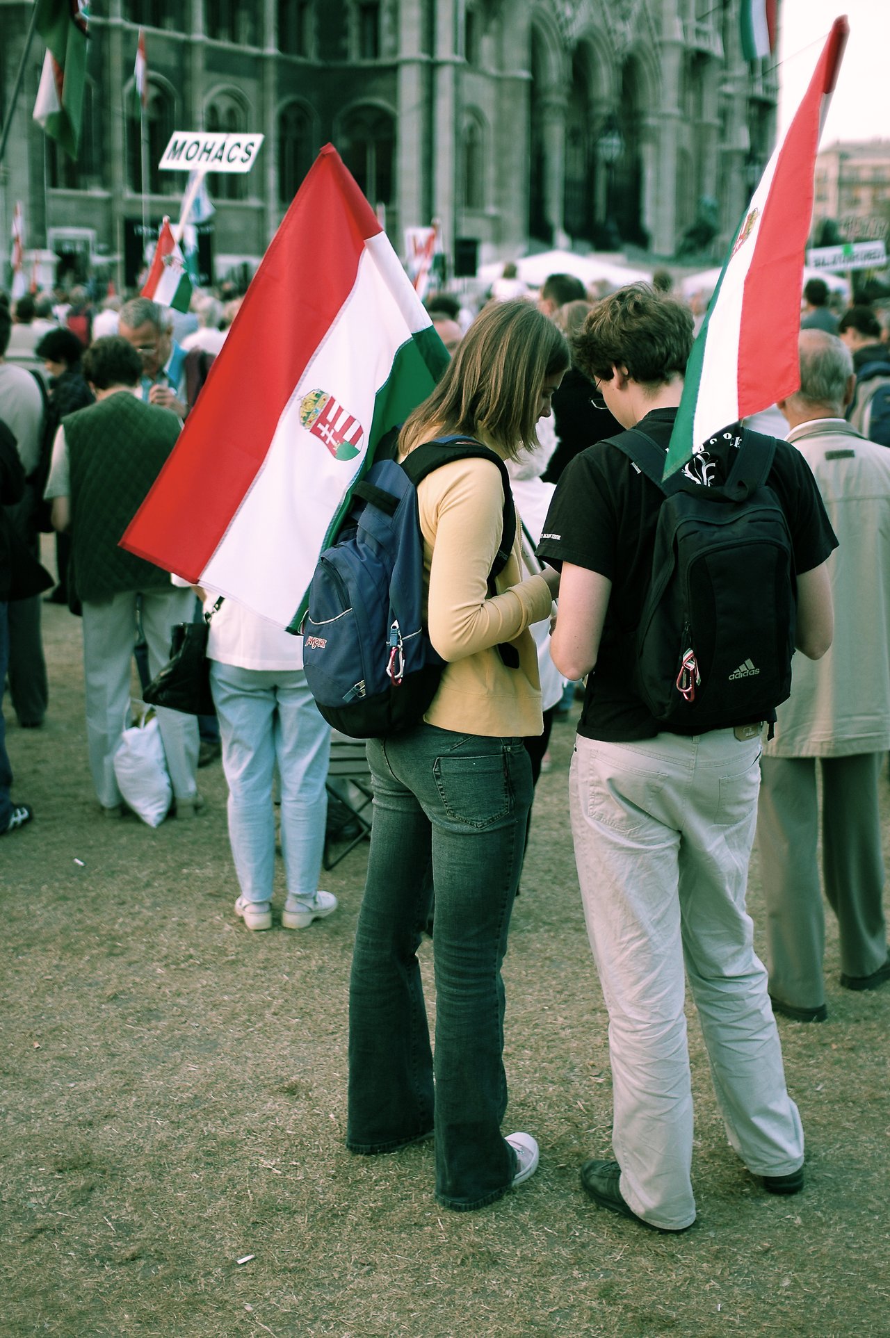 Two people with backpacks stand close together, holding Hungarian flags, during a peaceful protest with a crowd behind them.