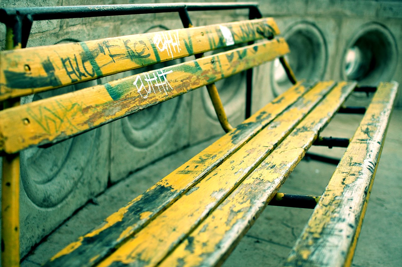 A worn yellow bench with graffiti on the wooden slats, placed against a concrete wall with circular openings.