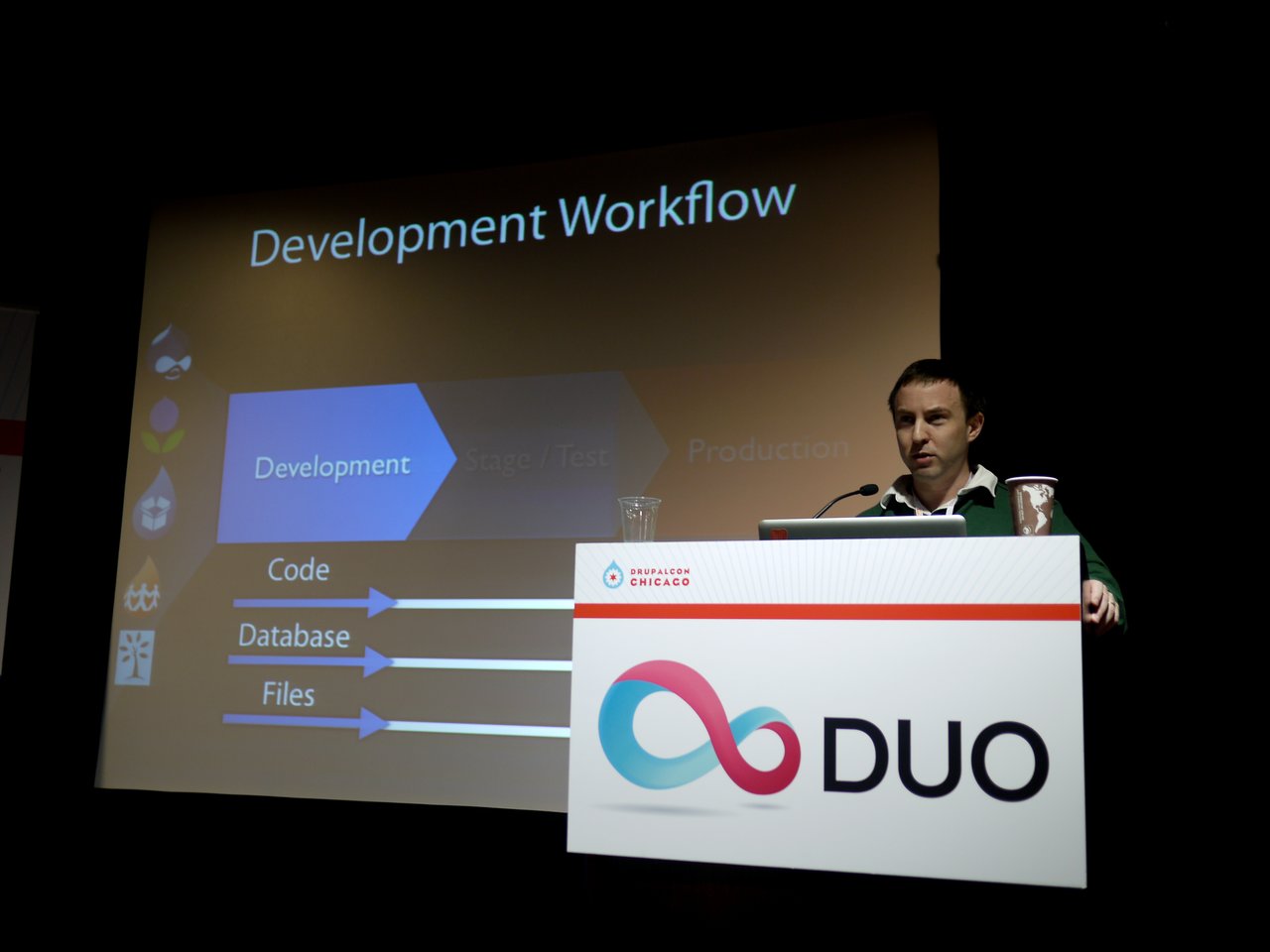 A speaker presents the Acquia DevCloud launch at DrupalCon Chicago 2011, discussing the development workflow on a large screen.