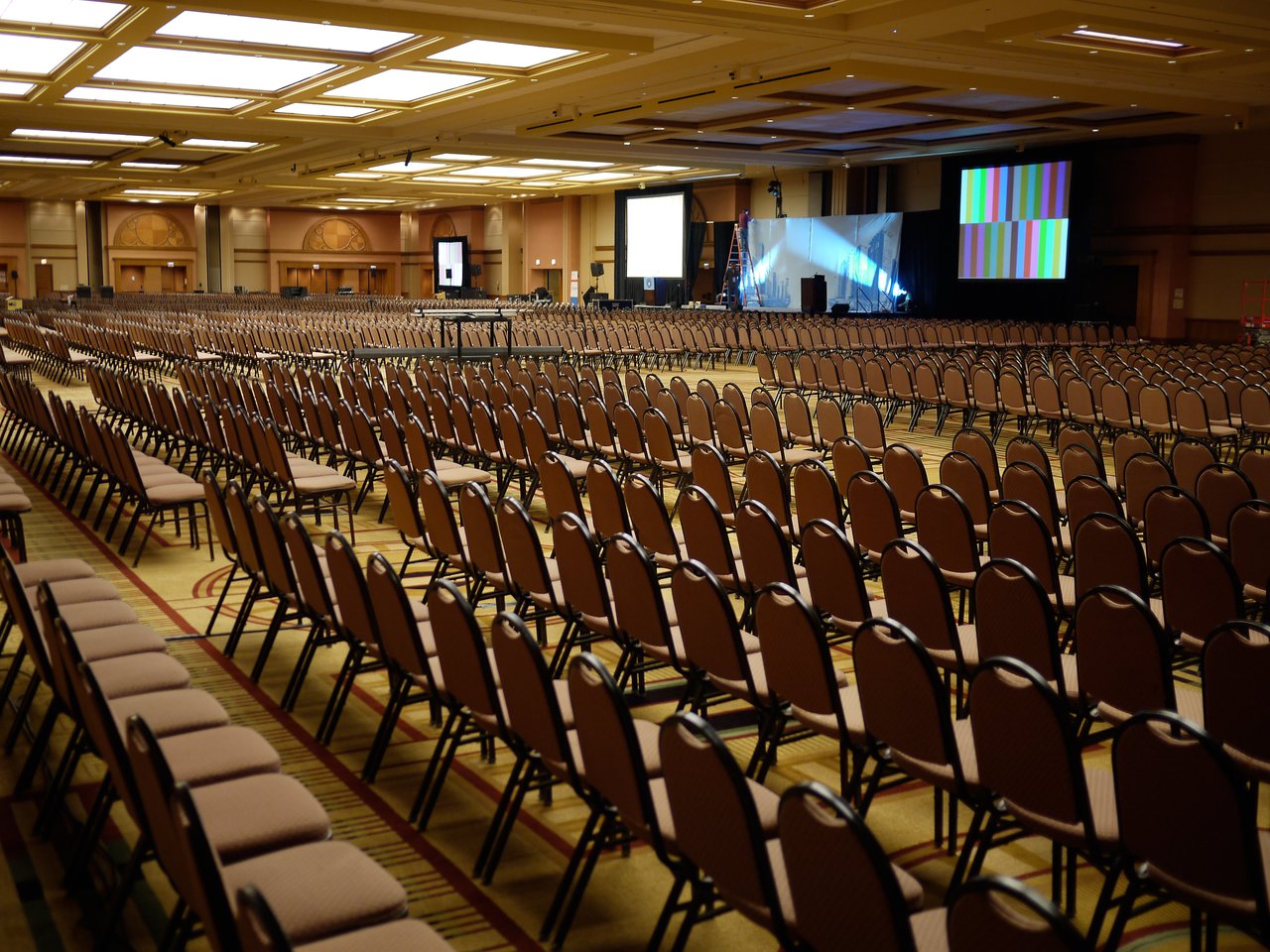 Large conference room with rows of empty chairs facing a stage with screens and lighting setup.