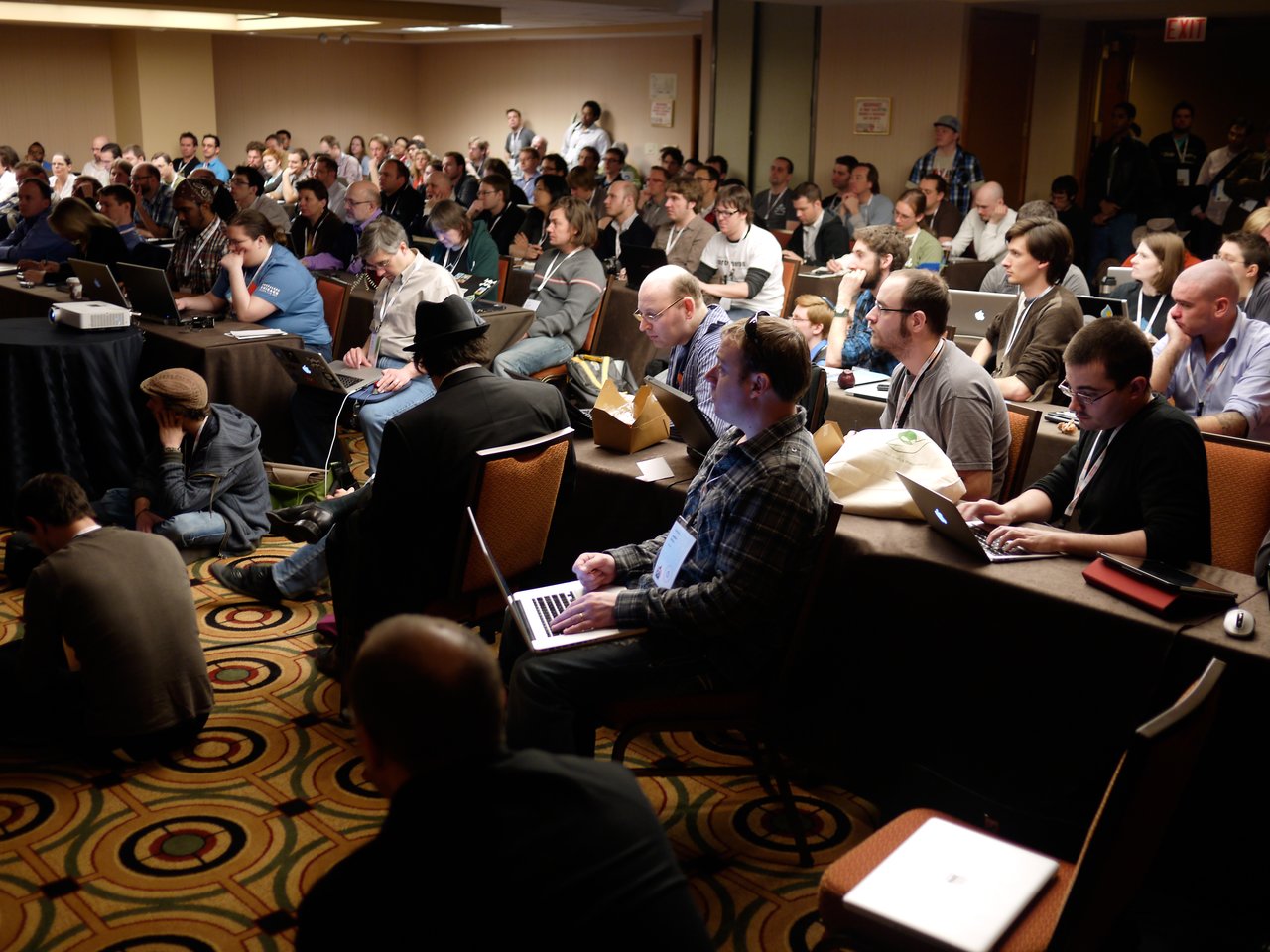 A large group of people attentively listening and taking notes on laptops during a DrupalCon core conversation session.