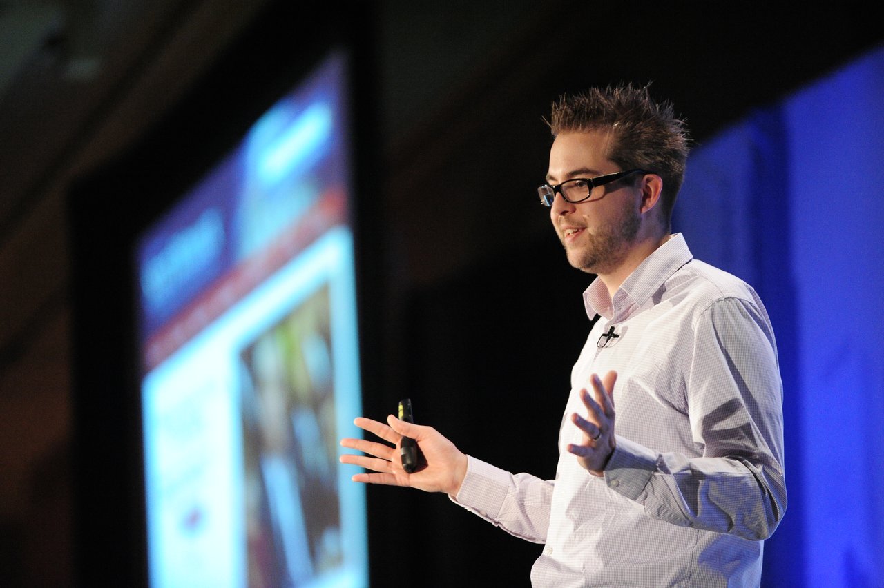A speaker in a white shirt and glasses gives a keynote presentation on stage, gesturing with both hands.