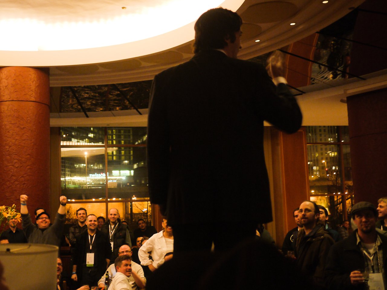 A man in a suit gestures while speaking to a cheering crowd in a dimly lit indoor setting.