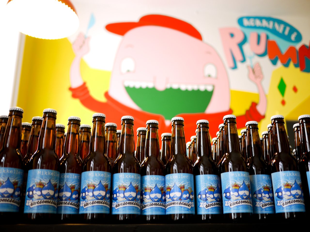 Rows of brown beer bottles with blue "Awesomesauce" labels, displayed in front of a colorful cartoon mural.